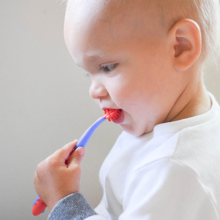 A young child with light hair in a white shirt practices dental hygiene, looking down while brushing their teeth with the 4-Stage Oral Care System baby toothbrush.