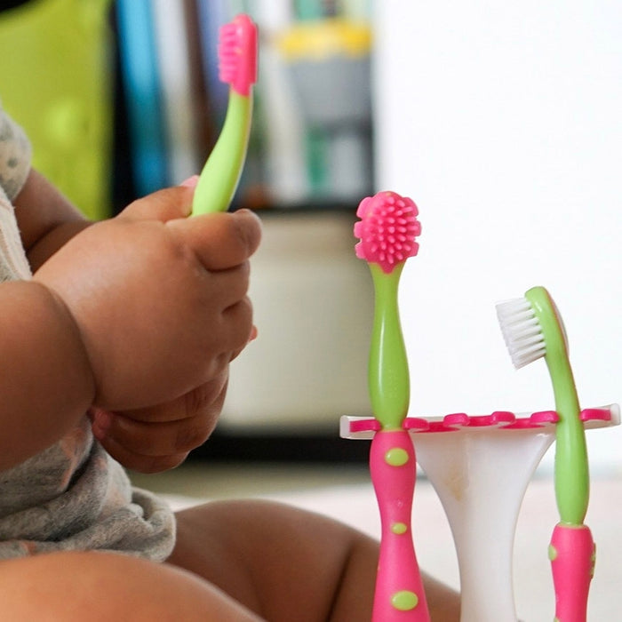 A baby holds a 4-Stage Oral Care System toothbrush, with other colorful baby toothbrushes in a stand nearby. The baby is sitting in a light-colored outfit, encouraging early dental hygiene habits.