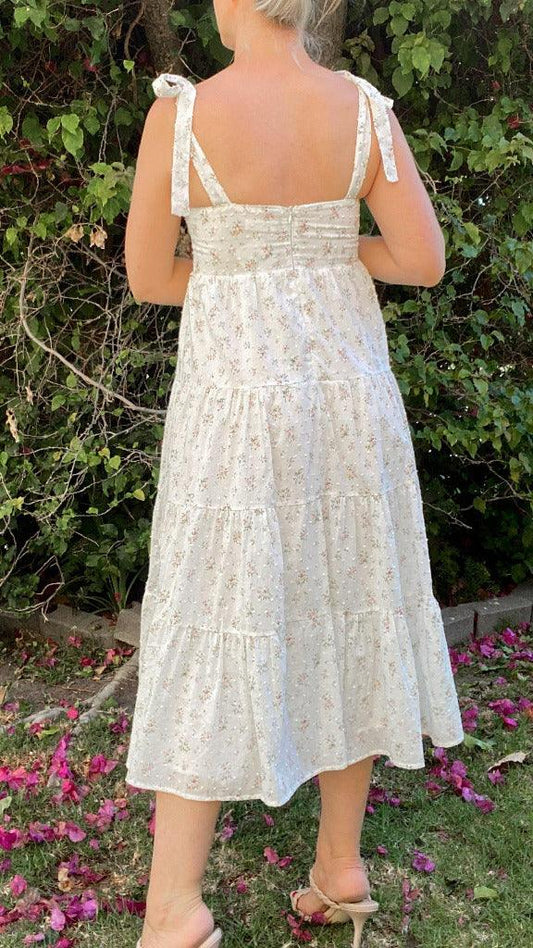 Back view of woman wearing a white floral self-tie tiered midi dress in a garden with green foliage