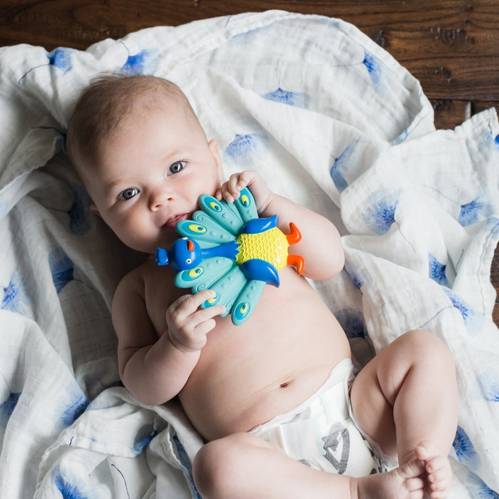 A baby in a diaper lies on a white blanket with blue patterns, holding and chewing on the Squeezy Teethe Soft Rubber Teether.