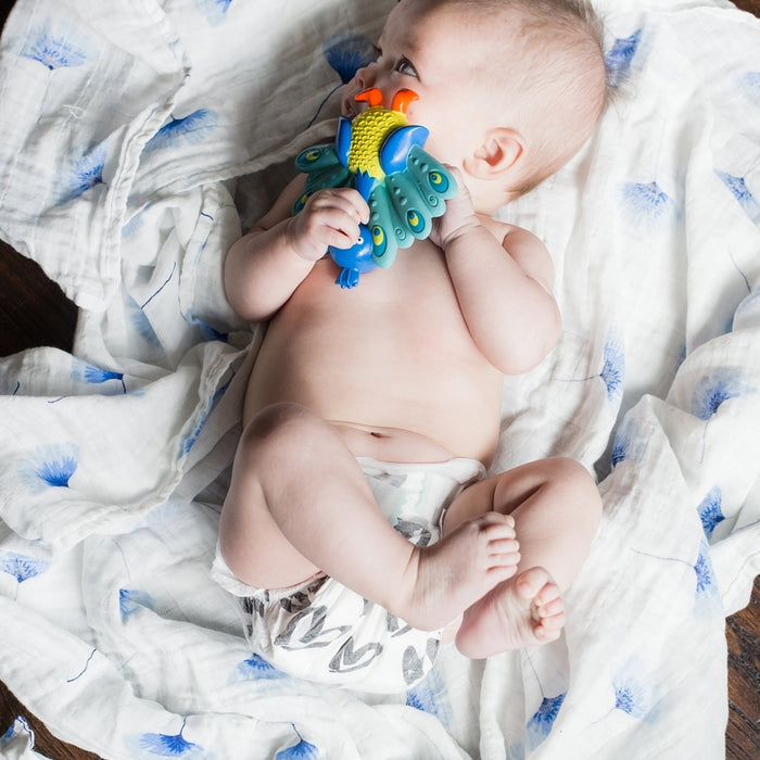 A baby in a diaper lies on a white blanket with blue patterns, holding and chewing on the Squeezy Teethe Soft Rubber Teether shaped like a colorful bird.